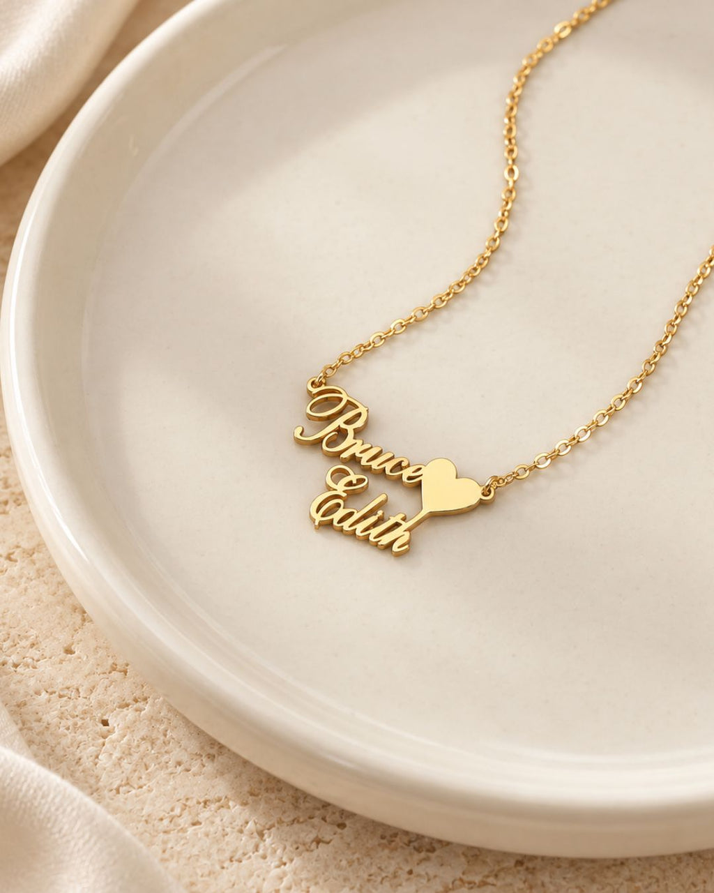 Collier doré personnalisé avec les prénoms Bruce et Edith reliés par un cœur, présenté sur une coupelle beige.