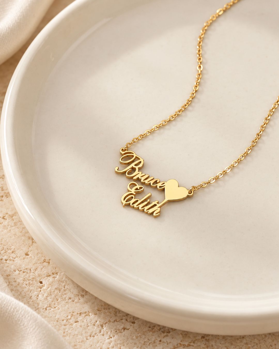 Collier doré personnalisé avec les prénoms Bruce et Edith reliés par un cœur, présenté sur une coupelle beige.