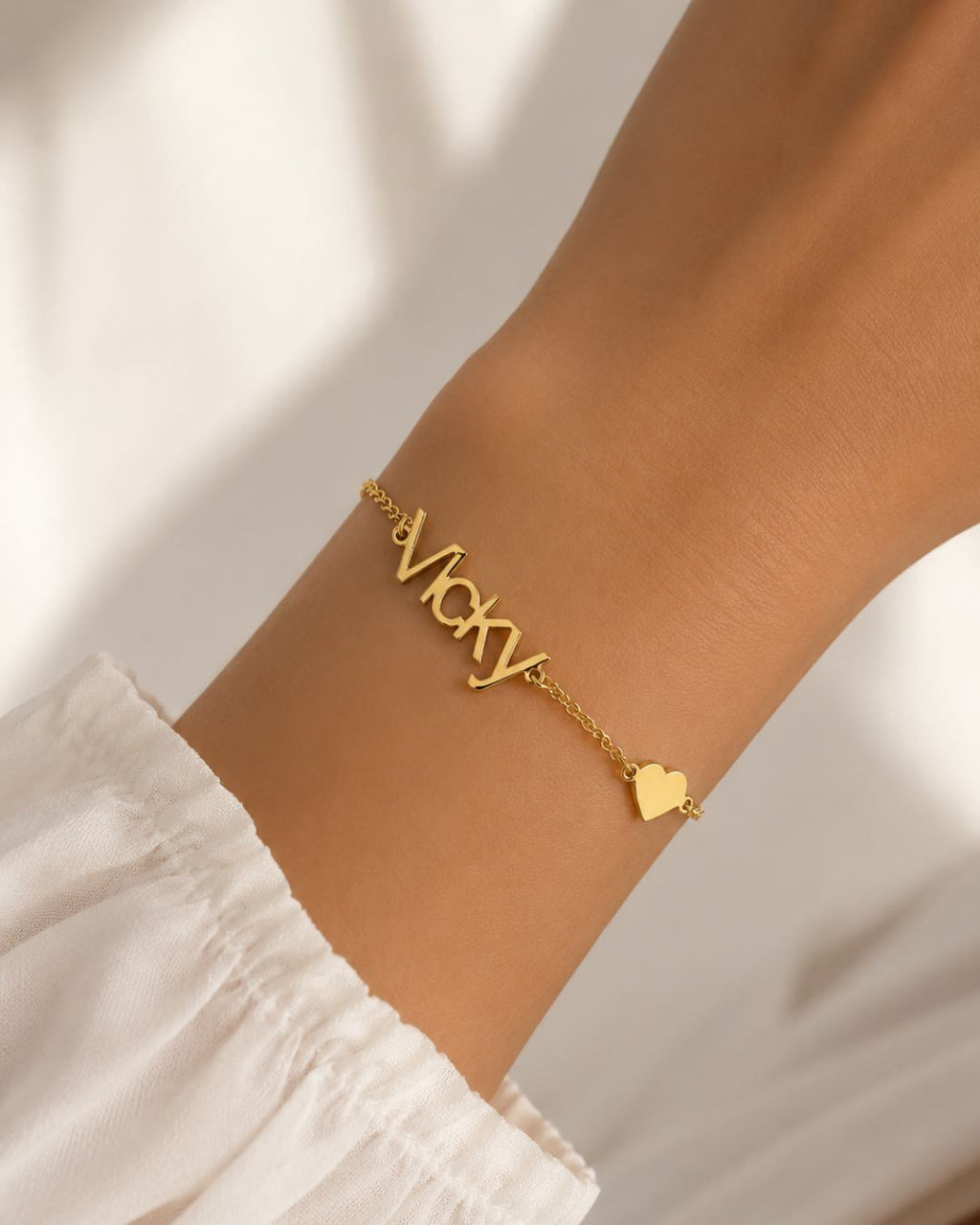Gold-finish Fidji Love personalised heart bracelet worn on a wrist beside a white sleeve.