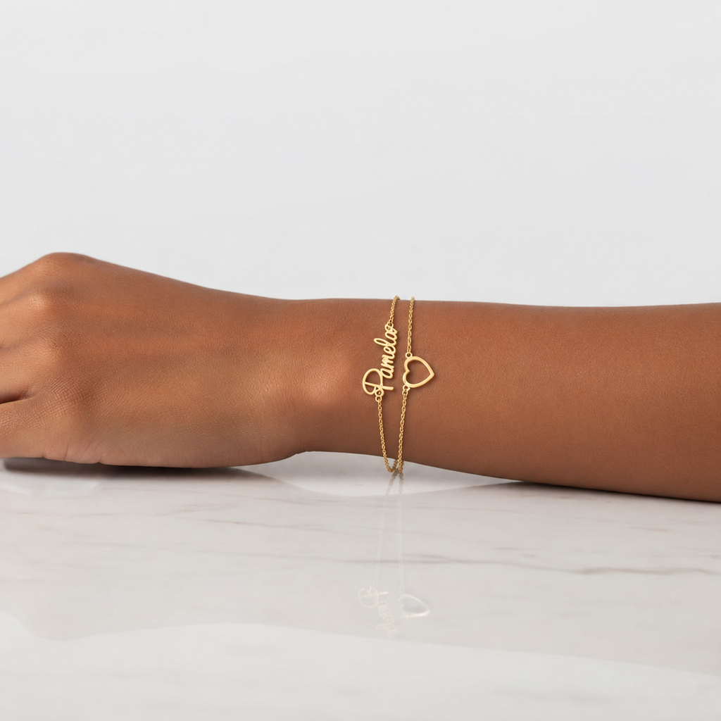 Gold-finish Hope double chain heart bracelet with “Pamela” lettering, worn on a wrist resting on white marble.