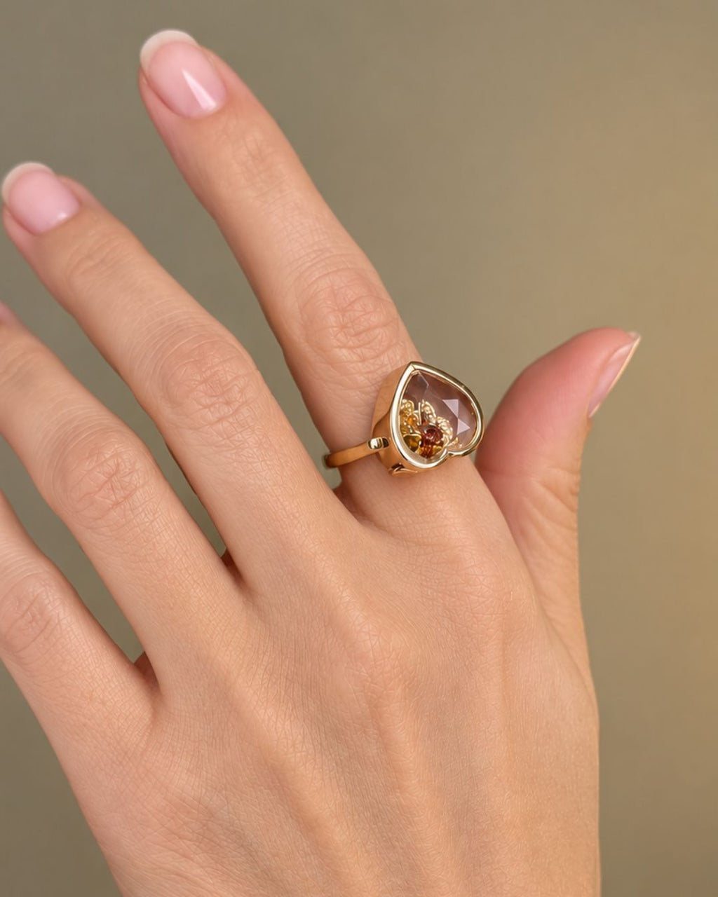 Bague Remember Ring dorée au cœur transparent contenant des charms, portée sur une main aux ongles roses.