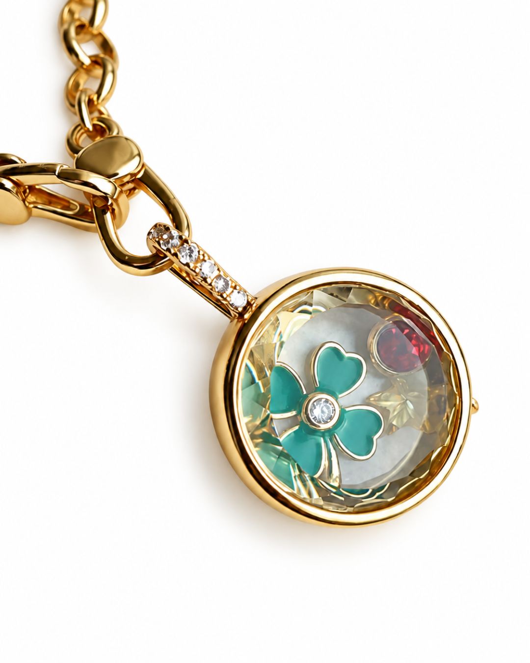 Close-up of a round gold-tone glass locket with a turquoise four-leaf clover charm and a red gemstone charm on a white background.