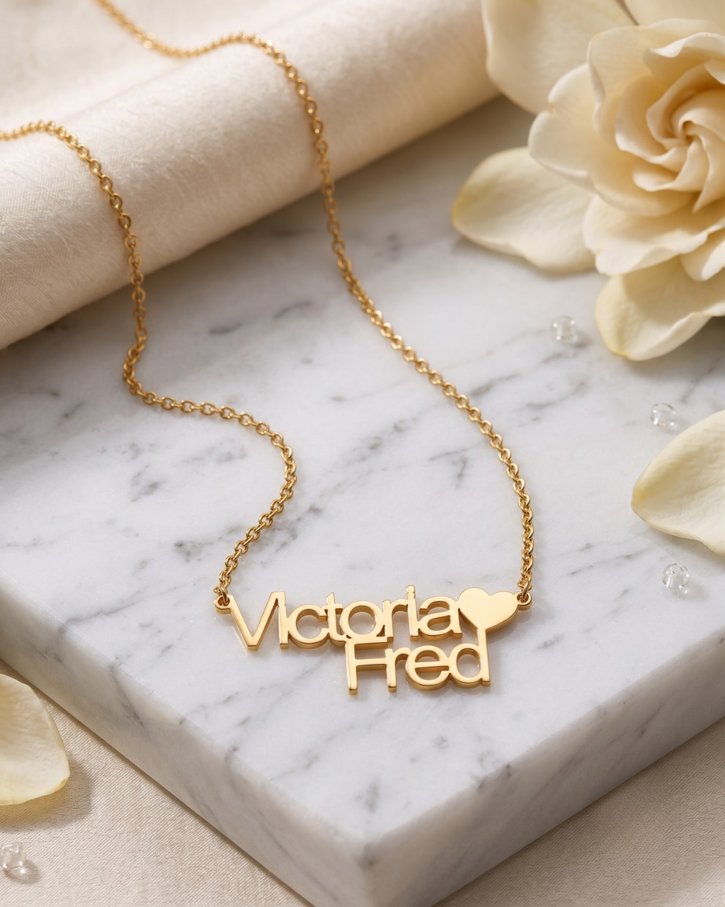 Gold-finish Together necklace with “Victoria” and “Fred” lettering and a heart detail, displayed with cream fabric and white flowers.