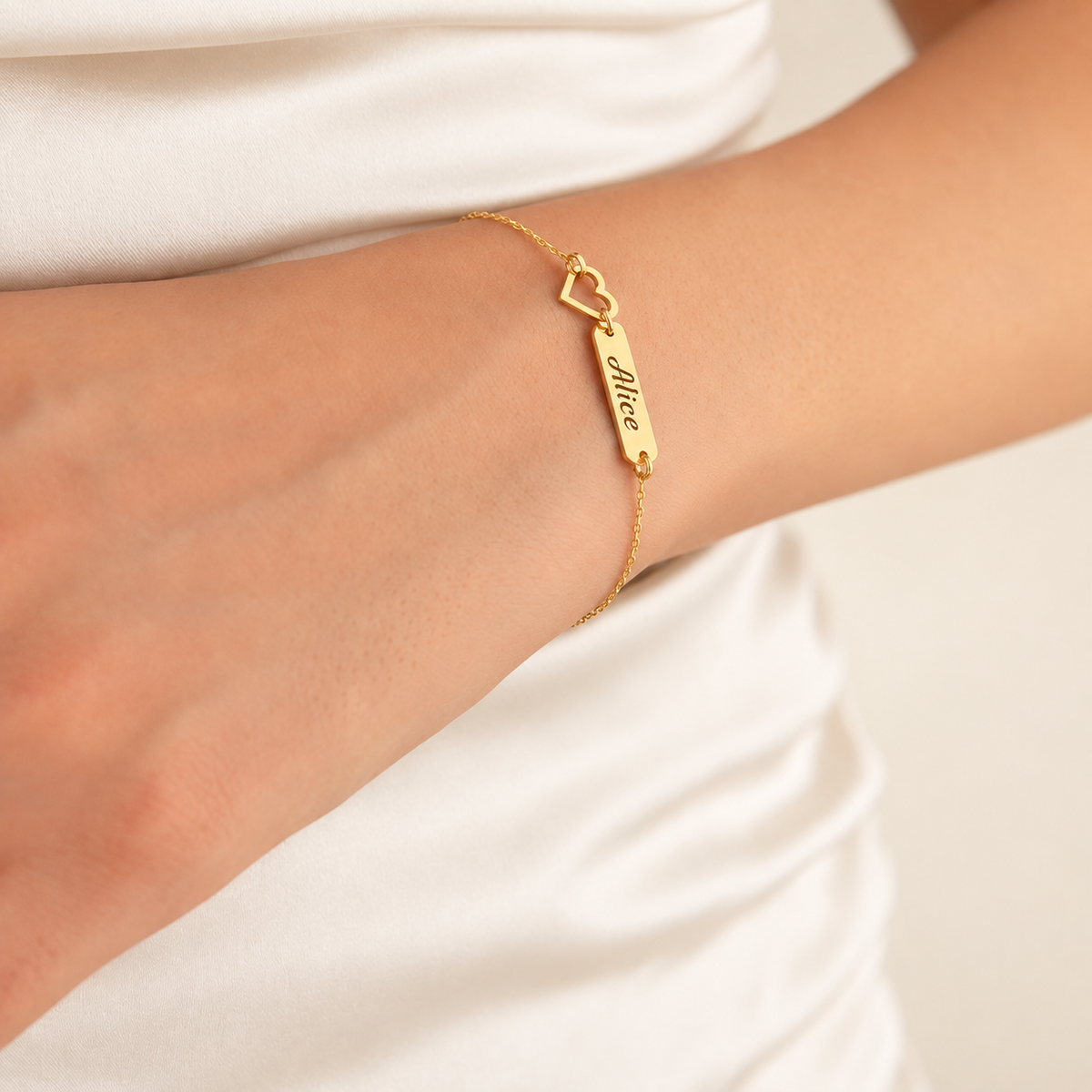 Gold-finish Venice personalised bracelet with “Alice” engraved on a rectangular bar and an open-heart detail, worn on a wrist against ivory fabric.